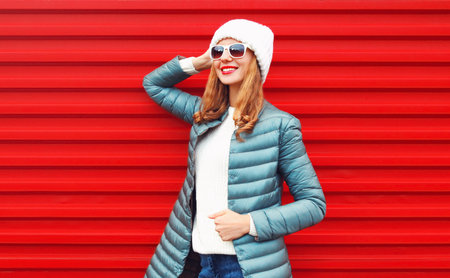 Portrait of happy smiling young woman posing wearing a jacket and white hat on red backgroundの写真素材