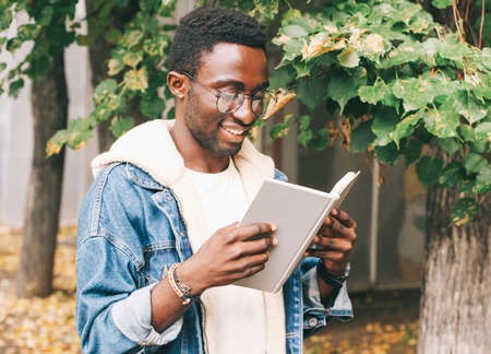 Portrait of young african man student reading a book wearing an eyeglasses in autumn city parkの写真素材