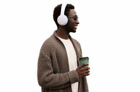 Portrait of stylish african man in wireless headphones listening to music with coffee cup looking away wearing brown knitted cardigan isolated on white backgroundの写真素材