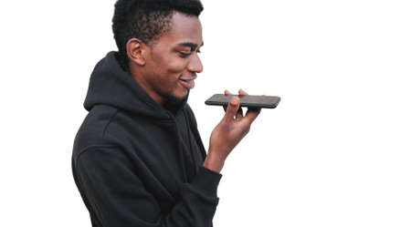 Portrait of modern young african man holding smartphone using voice command recorder, assistant or takes calling, looking at phone isolated on white backgroundの写真素材