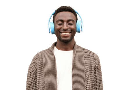 Portrait of happy smiling african man listening to music in wireless headphones wearing brown knitted cardigan isolated on white backgroundの写真素材