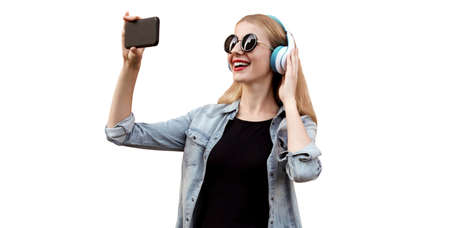 Portrait of happy smiling young woman taking selfie picture by smartphone in wireless headphones listening to music isolated on white backgroundの写真素材