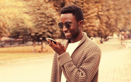 Portrait of happy smiling african man with smartphone using voice command recorder, assistant or takes calling in autumn city parkの写真素材