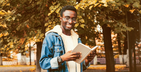 Portrait of happy smiling young african man student reading a book wearing eyeglasses in autumn city...