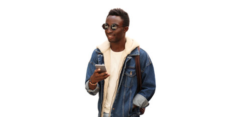 Portrait of modern young african man with smartphone wearing jean jacket, sunglasses and backpack isolated on white backgroundの写真素材