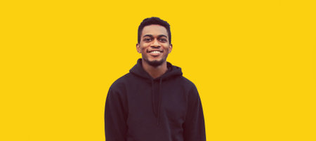 Portrait of modern smiling young african man looking at camera wearing black hoodie isolated on yellow backgroundの写真素材