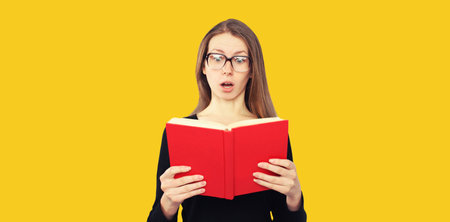 Portrait of shocked student girl in eyeglasses looking in the opened book on yellow backgroundの写真素材
