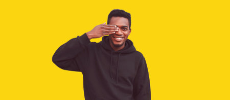 Portrait of happy smiling african man covering his face with his hand and looking by one eye wearing black hoodie isolated on yellow backgroundの写真素材