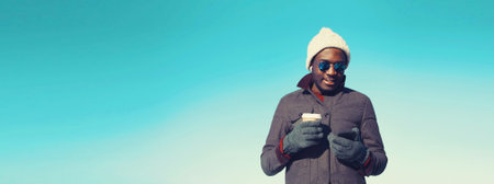 Portrait of young african man with smartphone wearing winter hat, jacket on blue sky background, blank copy space for advertising textの写真素材