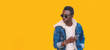 Portrait of stylish young african man with smartphone looking away isolated on yellow background, blank copy space for advertising textの写真素材