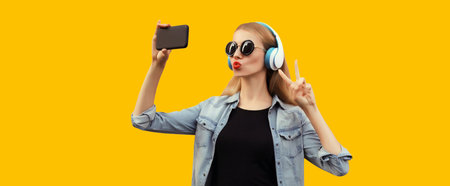 Portrait of stylish young woman taking selfie with smartphone in wireless headphones listening to...