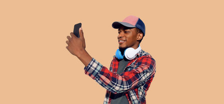 Portrait of modern smiling young african man taking selfie with smartphone listening to music in headphones wearing baseball cap isolated on brown backgroundの写真素材