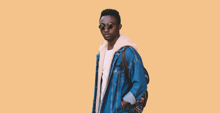 Portrait of stylish young african man model wearing denim jacket, backpack isolated on brown...