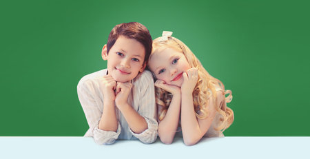 Portrait of happy cute smiling two children pupils sitting at the desk table together on green school chalk board backgroundの写真素材