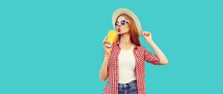 Portrait of young woman drinking fresh juice wearing summer straw hat, checkered shirt isolated on blue backgroundの写真素材