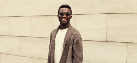 Portrait of stylish smiling young african man wearing brown knitted cardigan on city street backgroundの写真素材