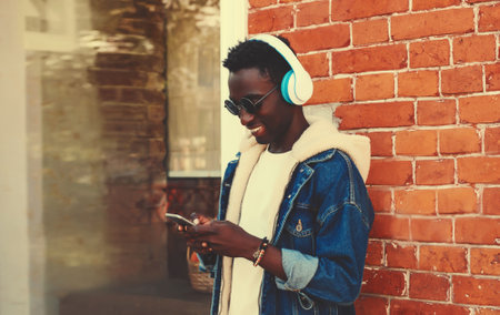 Portrait of stylish young african man with phone listening to music in wireless headphones on city streetの写真素材