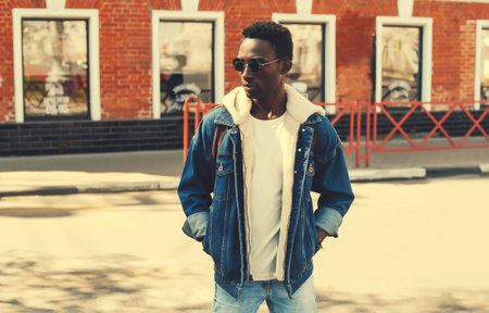 Portrait of stylish young african man posing wearing denim jacket, backpack on city street backgroundの写真素材