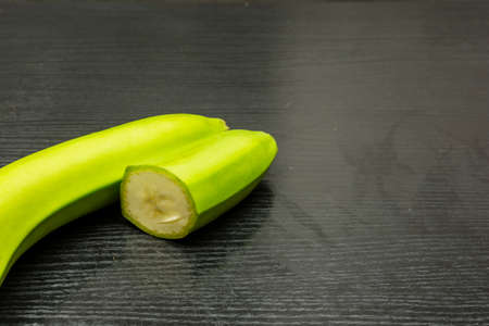 A green banana cut in half lies on a dark wooden table.の写真素材