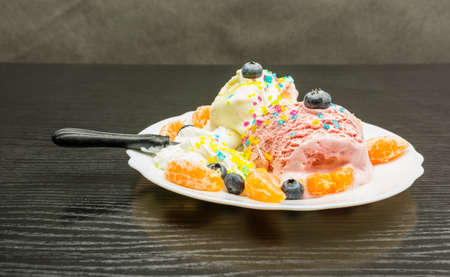 A frosty dessert on a plate with a dipped spoon - ice cream, whipped cream, fresh fruit and neon sugar decorations on a wooden table.の写真素材