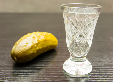 Vodka in a crystal shot and pickled cucumber on a wooden dark basis.