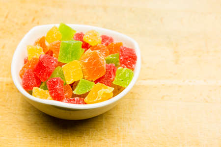Colorful cubes of candy pineapple in a white bowl set on a wooden substrate.の写真素材