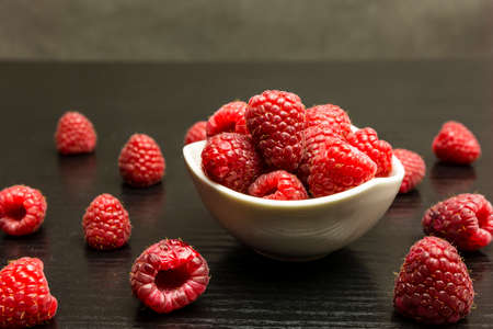 Ripe raspberries in a white bowl and on a dark table.の写真素材