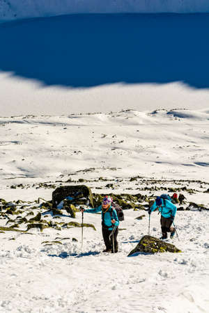 Bukowina Tatrzanska, Poland - January 12, 2020: Two women (tourists) in turquoise jackets and trekking poles while approaching a steep snowy slope in the mountains. Winter mountain tourism.のeditorial素材