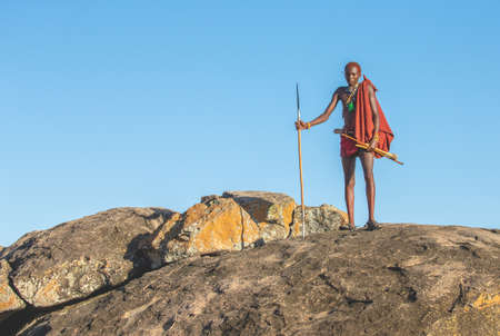 ANZANIA, EAST AFRICA - AUGUST 12, 2018: Young Masai warrior is standing on a big stone in traditional clothing with a spear in the savannah against a blue sky. Tanzania, East Africa, August 12, 2018.のeditorial素材