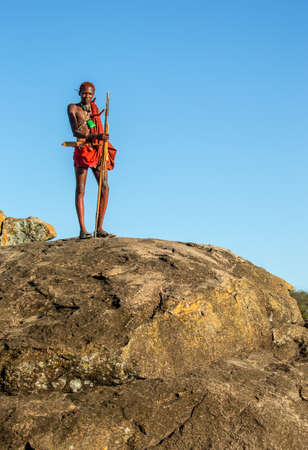 ANZANIA, EAST AFRICA - AUGUST 12, 2018: Young Masai warrior is standing on a big stone in traditional clothing with a spear in the savannah against a blue sky. Tanzania, East Africa, August 12, 2018.のeditorial素材