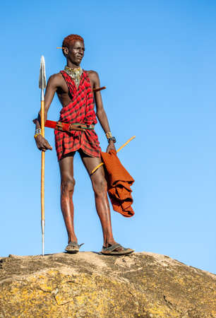 ANZANIA, EAST AFRICA - AUGUST 12, 2018: Young Masai warrior is standing on a big stone in traditional clothing with a spear in the savannah against a blue sky. Tanzania, East Africa, August 12, 2018.のeditorial素材