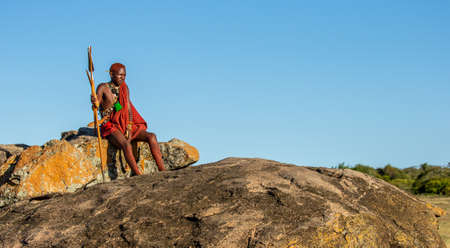 Young Masai warrior is sitting on a big stone in traditional clothing with a spear in the savannah against a blue sky. Tanzania, East Africa, August 12, 2018.のeditorial素材