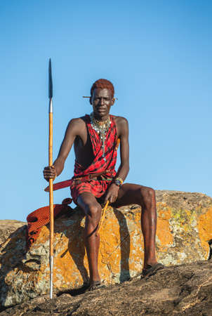Young Masai warrior is sitting on a big stone in traditional clothing with a spear in the savannah against a blue sky. Tanzania, East Africa, August 12, 2018.のeditorial素材