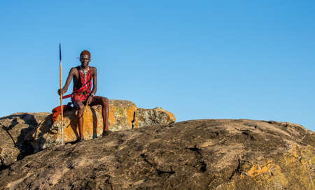 Young Masai warrior is sitting on a big stone in traditional clothing with a spear in the savannah against a blue sky. Tanzania, East Africa, August 12, 2018.のeditorial素材