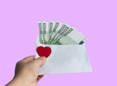 A hand holds a paper envelope with 100 euro banknotes and a red heart on a pink background. close-up. Conceptual gift for Valentine's Day, wedding, birthday.の写真素材