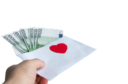A hand holds a paper envelope with 100 euro banknotes and a red heart on a white background. Conceptual gift for Valentine's Day, wedding, birthday. close-up.の写真素材