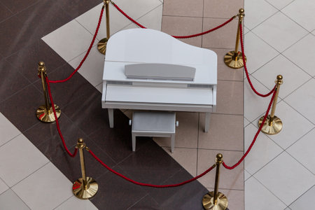 A white grand piano on a ceramic tiled floor is fenced with golden poles and red ropes. view from above.の写真素材