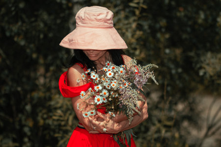 A girl in a pink hat, glasses in a red dress holds a bouquet of wild flowers in her hands on a...