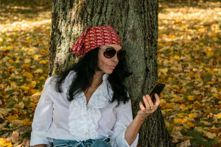 A girl with glasses sits under a tree among yellow foliage and calls on the phone. autumn mood.の写真素材
