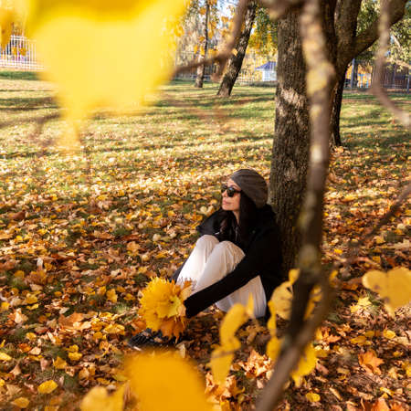 A girl sits in an autumn park and holds yellow maple leaves in her hand. Yellow blurred leaves in the foreground. autumn mood.の写真素材