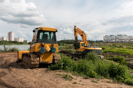 A caterpillar excavator digs the earth with a bucket, and a bulldozer clears the earth with a metal shield. Clearing the construction site. Drainage of swamps. Peat mining.の写真素材