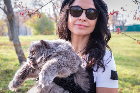 The girl in glasses holds a cat in her arms against a background of yellow autumn leaves and red rowan. autumn mood. Walk with the cat.の写真素材