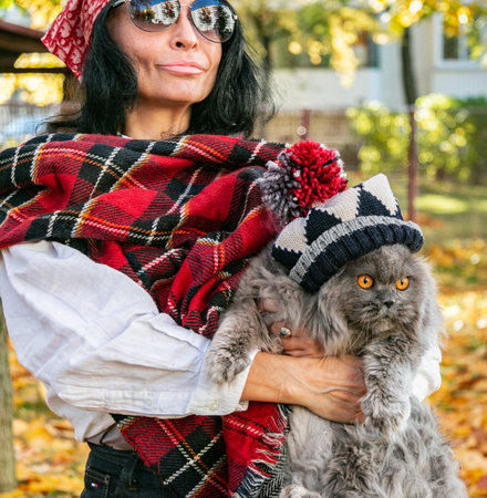 A girl in sunglasses with a scarf on her head holds a British cat in a hat with a pompom in her arms against the background of yellow autumn maple leaves. Walk with a cat. autumn mood. close-up.の写真素材