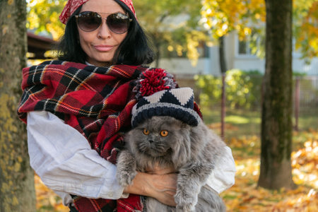 A girl in sunglasses with a scarf on her head holds a British cat in a hat with a pompom in her arms against the background of yellow autumn maple leaves. Walk with a cat. autumn mood. close-up.の写真素材
