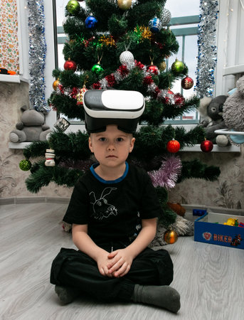 Minsk. Belarus. 01/12/2022. A little boy with virtual reality glasses sits near a Christmas tree with toys.のeditorial素材