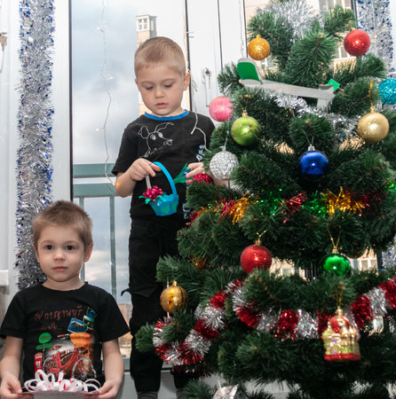 Minsk. Belarus. 01/12/2022. Excited little brothers celebrate New Year and Christmas at home. Two happy little boys near the Christmas tree with toys. The boys are holding handmade Christmas gifts.のeditorial素材