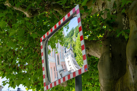 A rectangular traffic mirror with a red and white frame and a distorted reflection of a city. The mirror is mounted in front of a tree with green leaves and the sun is shining.の写真素材