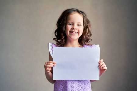curly haired girl holding a white sheet of paper smiling on a gray background.の写真素材