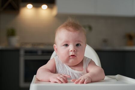 a small child in a white bodysuit sits in a high chair in the kitchen and waits to eat.の写真素材