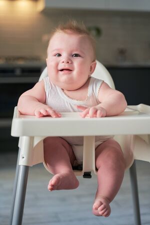 a small baby is sitting in a child's chair in the kitchen and waiting to eat.の写真素材